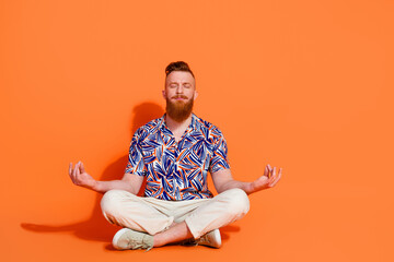 Full length photo of focused peaceful guy wear print shirt white trousers sit in meditation pose isolated on orange color background