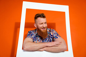 Photo of optimistic guy with red beard wear print shirt hold hands crossed in frame blinking eye isolated on orange color background