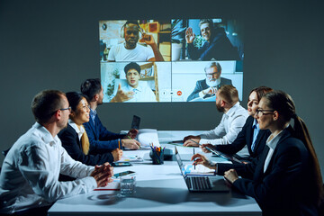Employees sit in modern office boardroom, discussing projects and documents over a video call with...