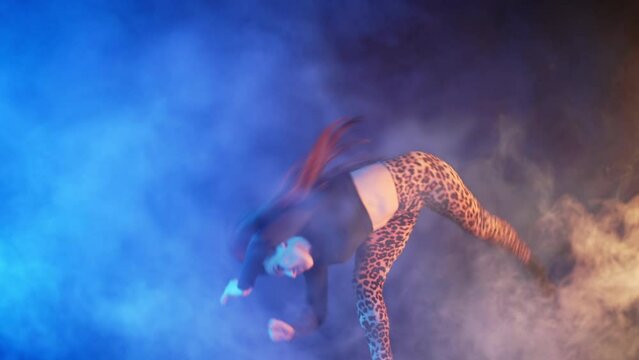 Woman gymnast making twist, demonstrates extreme trick in smoke neon studio. Female acrobat jumping front flip. Tricking, parkour, jumping practice concept