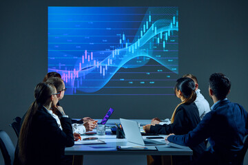 Office employees gather in boardroom, analyzing digital stock chart projected on screen during...