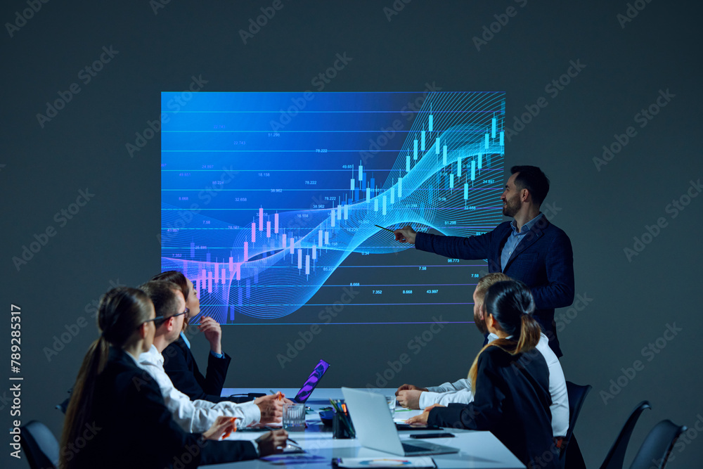 Wall mural team members sitting in boardroom setting, reviewing digital stock chart on screen as part of a busi