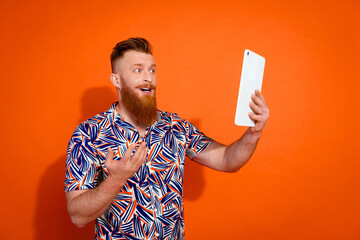 Photo of handsome brutal guy with red beard wear print shirt look at tablet talk on selfie camera isolated on orange color background