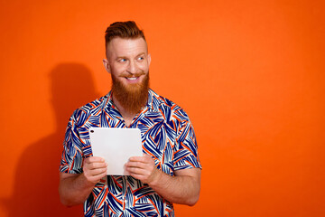 Photo of nice guy with red beard wear print shirt hold tablet look at discount empty space isolated on vivid orange color background
