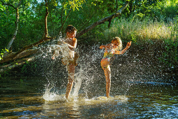 children playing in the river. A girl and boy raises her hands up in the water and splashes water drops - obrazy, fototapety, plakaty