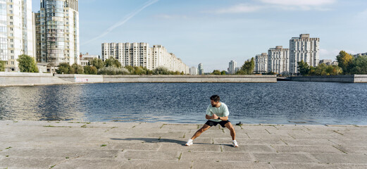 A man stretches in a side lunge along a spacious riverbank promenade with a backdrop of city...