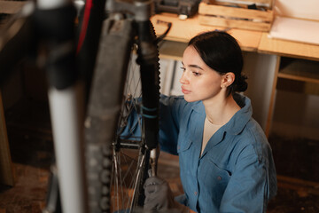 Fototapeta na wymiar Beautiful brunette long haired female worker crouching and repairing bicycle in garage. Bike workshop interior.