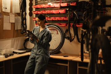 Young professional mechanic woman in blue overalls use mobile phone browsing fix problem in bike...