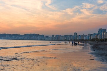 People walking on the beach near the sea during sunset in the city of Santos, Brazil. Waterfront buildings in the background.