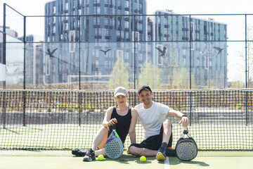 Portrait of positive young woman and adult man standing on padel tennis court, holding racket and ball, smiling 