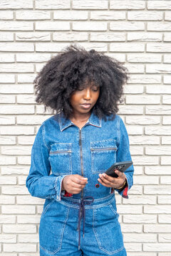 Against a white brick wall, a young Black woman with afro-textured hair is focused on her smartphone. Her denim jumpsuit suggests a casual, modern vibe, while her absorbed expression indicates