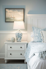 A white nightstand in front of the bed, light blue and grey wainscoting, soft neutral colors, vintage inspired, coastal style, soft lighting, with art on wall above headboard.