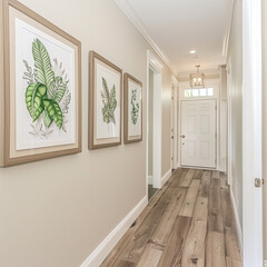 Three posters with wooden frames hang on a beige wall in a narrow corridor with a doorway and front door, a chandelier on a white ceiling, and a wooden floor.