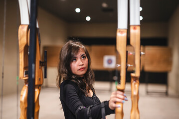 An Asian woman examines and chooses a bow at an indoor archery range, showcasing a casual yet...
