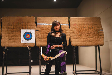 An amateur archer, a young Asian woman, is seen at an indoor archery range, equipped with a bow and...
