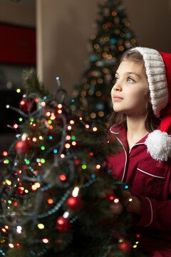 Happy girl of 7 years old in red pyjamas and Santa Claus hat looks out of window next to Christmas tree with side of garlands with bokeh