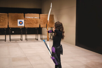 A young Asian woman is pictured practicing archery, focusing intently on the target at an indoor range. View from behind of enthusiast aiming for the bullseye. - obrazy, fototapety, plakaty