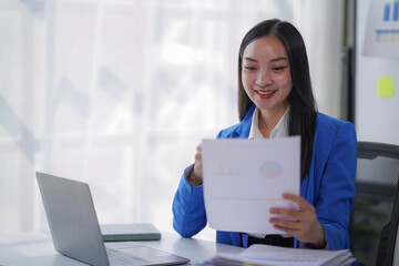 Asian businesswoman at desk smiling as she reviews financial reports and charts on laptop, analyzing data and paperwork to plan strategy, track performance and drive growth