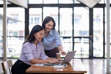Fototapeta premium Two smiling businesswomen collaborating in a modern office environment, reviewing a document and using a laptop.
