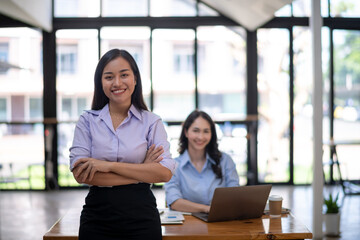 A confident female leader stands arms crossed with her teammate working on a laptop in the background in a sunny office.