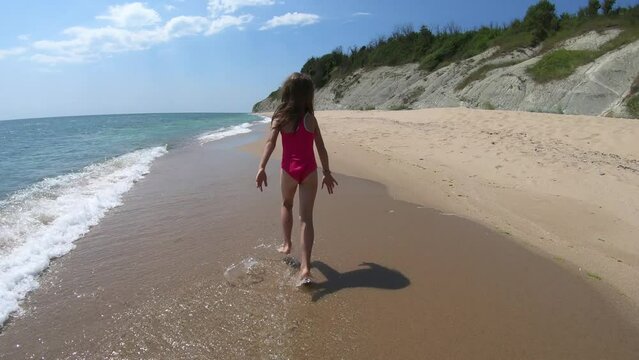 Happy playful young girl, kid running barefoot in pink swimwear on sandy beach in summer vacation, sea waves approaching the shore