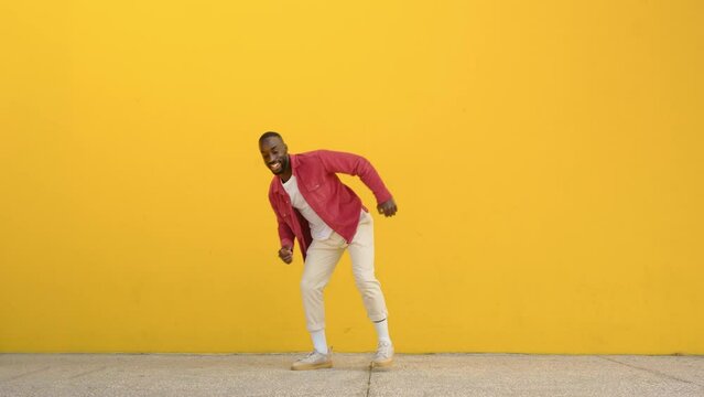Happy active energetic crazy gen z young Black man jumping at yellow urban wall. Excited cool funky African guy having fun dancing moving on background celebrating victory. Full body video.