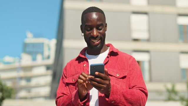 Happy young African Black man hipster standing on city street outdoors holding smartphone using mobile apps looking at cell phone technology device chatting in social media, dating applications.