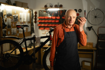 Old man male bicycle mechanic listening to voice message while standing in garage or bicycle repair...