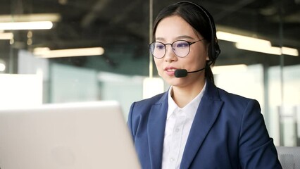 Asian female in wireless headset customer service operator talking on video call on laptop sitting in business office. Agent of the call center advises the client, answering questions. Close up
