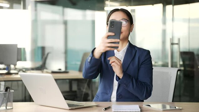Frustrated asian businesswoman complains about lack of Wi-Fi signal using mobile phone sitting at workplace in business office. Worried puzzled woman worker is dissatisfied with the work of smartphone