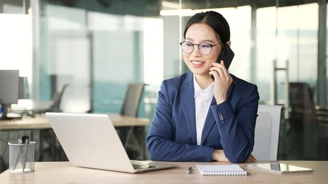 Confident young asian businesswoman talking on the phone while sitting at workplace in business office. Busy woman in formal suit discusses business with a partner or has a conversation with a client