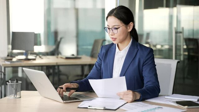 Busy young asian businesswoman female accountant doing paperwork using laptop computer sitting at workplace in business office. Woman financier in glasses, making financial report or writing tax form