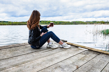 Woman with mobile phone and coffee sitting on wooden pier bridge near by lake.