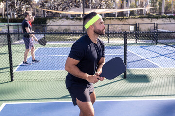 Pickleball player wearing a head band for sweat holding a paddle. He is wearing a black shirt and is ready for the ball to come his way on the outdoor court. 