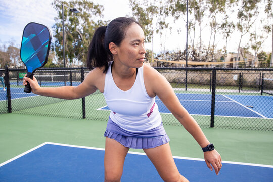 A pickleball player holds her paddle in anticipation of a serve coming at her. The woman is fit and having fun with outdoor exercise. The sport is popular and accessible. 