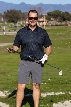 Adult male golfer wearing sunglasses and a glove stands and smiles for a portrait while hitting golf balls at a range. 