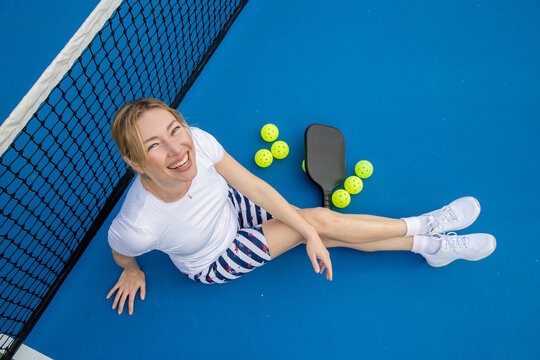 Female pickleball player sitting on the blue court leaning against the net with her paddle and balls on the ground next to her. She is looking up and smiling. 