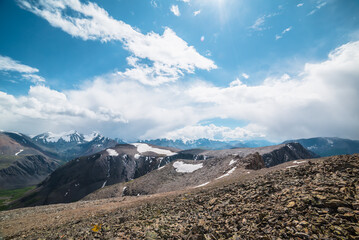 Awesome view to sunlit big stony ridge with snowfield and sheer crags against snow-covered mountain range with snow-capped peaked top. High rocky pass and large snowy pointy peak in clouds in blue sky - 789423323