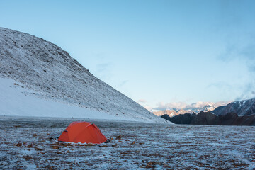 Orange tent on snow-covered stony pass. Snowy stone hill and rocky mountain ridge in sunlight at early morning. Red tent in high mountains in freshly fallen snow. Low clouds in blue sky at sunrise. - 789423329