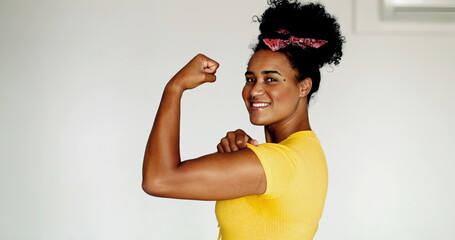 Happy Confident young African American woman showing strength, flexing arm looking at camera. One black latina shows muscle, we can do it symbol, wearing yellow shirt - obrazy, fototapety, plakaty