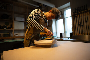 Sanding wood with orbital sander at workshop. Focused man carpenter polishes wooden seat of a...