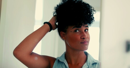 One happy young black Brazilian woman adjusting hairstyle in front of mirror getting ready to go out. Stylish African American 20s adult girl smiling feeling confident