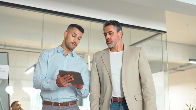 Busy professional executive business team of two men working together walking in office holding tab using digital tablet computer tech managing data talking going in corporate office hallway