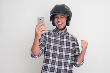 Adult Asian man wearing motorcycle helmet looking to his phone with excited gesture