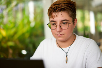 Transgender pro focuses laptop work in office. Inclusive environment, modern workplace diversity. Confident individual with glasses, casual attire. Background plants indicate eco-friendly space.