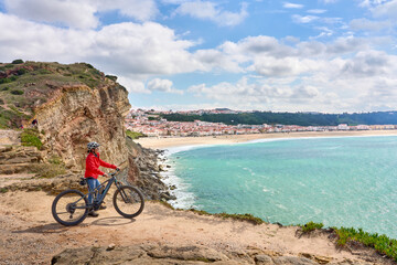 nice senior woman riding her electric mountain bike on the rocky cliffs above the city of Nazare at...