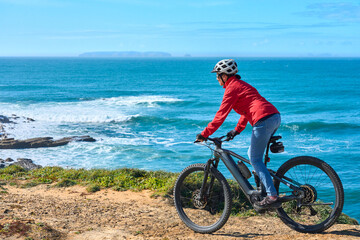 Brave senior woman riding her electric mountain bike on the rocky cliffs of Peniche at the western...