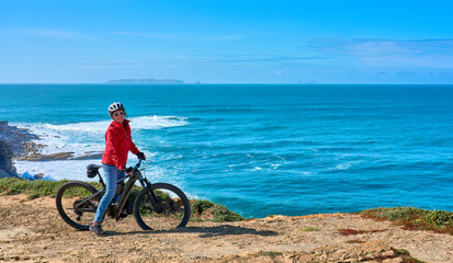 Fototapeta na wymiar Brave senior woman riding her electric mountain bike on the rocky cliffs of Peniche at the western atlantic coast of Portugal