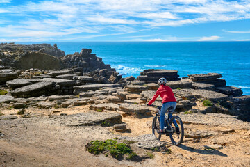 Brave senior woman riding her electric mountain bike on the rocky cliffs of Peniche at the western atlantic coast of Portugal - 789461173