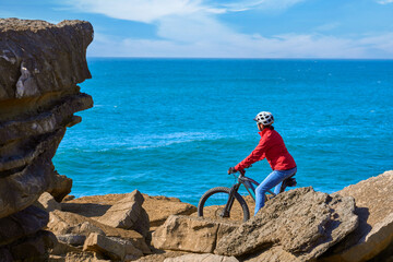 Brave senior woman riding her electric mountain bike on the rocky cliffs of Peniche at the western atlantic coast of Portugal - 789461574